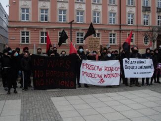 Na zdjęciu widac osoby na demonstracji, które pozują z transparentami i mają anarchistyczne, czare flagi.